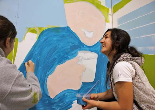 Logan Ball and Aubrey Romo of Aleknagik paint a base layer on Apayo Moore's mural, which will depict chuckling, storytelling elders with subsistence scenes in the background. (Photo by Hannah Colton/KDLG)