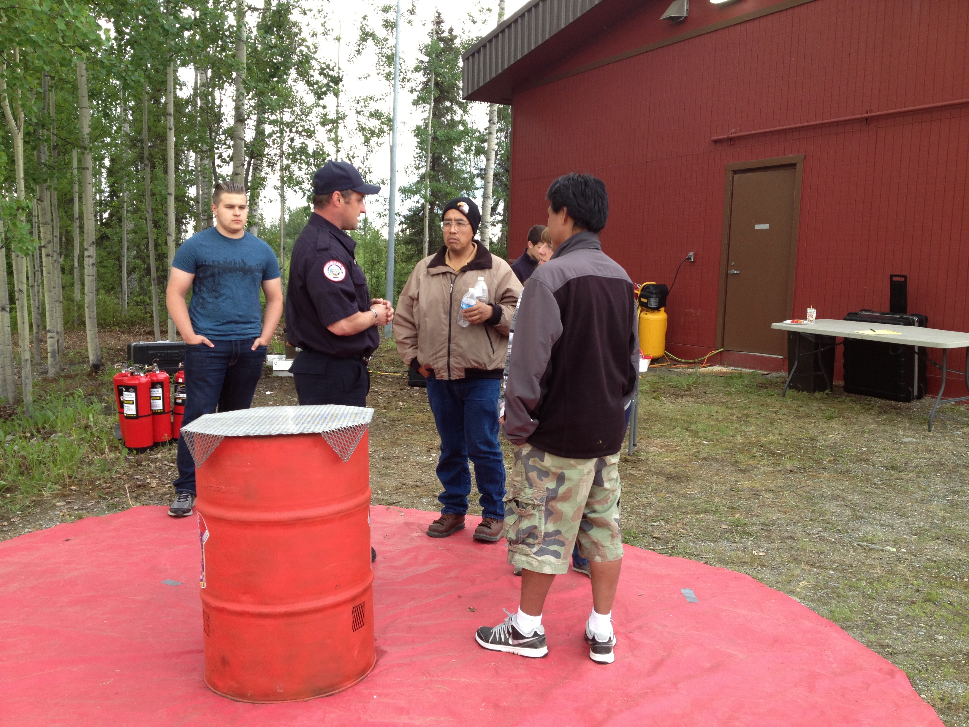  Zach Katzenberger, second from left, a fireman with the West Lakes fire department, exhibits a correct burn barrel setup to onlookers at Big Lake's fire safety event on Saturday. (Photo by Ellen Lockyer, Alaska Public Media - Anchorage)