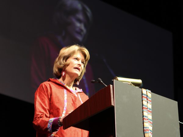 Woman at a podium in a red parky with her image projected behind her.