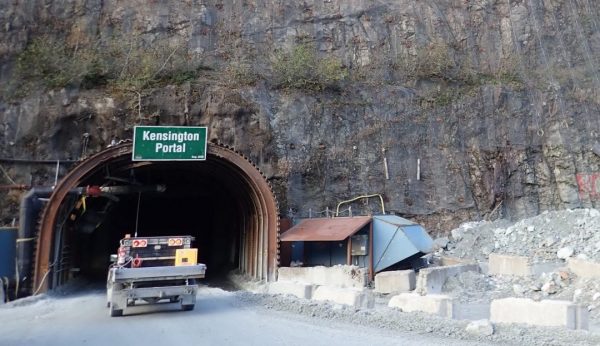 A truck enters a tunnel in a granite mountain. There is a green sign that says "Kensington Portal" above the tunnel and a blueish vent on the right side next to the vehicle. 