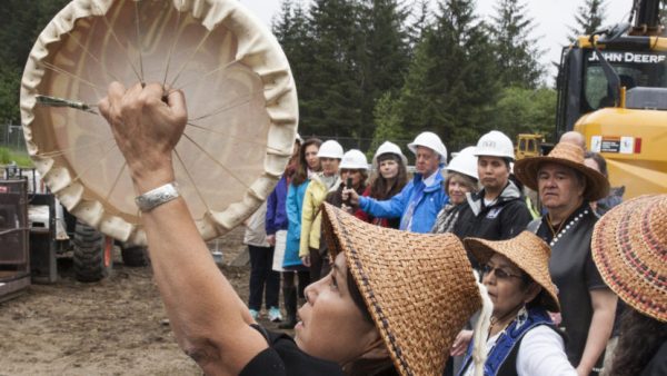A woman in a woven hat holds a skin drum in the air as people in hard hats look on. 
