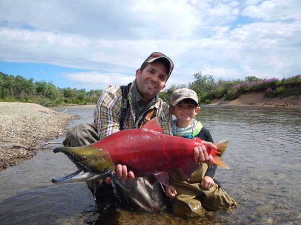 Donald Trump Jr. and his son in  river shallows. Trump jr. holds in front of him a sockeye salmon that is bright red with a green head.