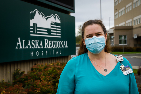 A woman stands in front of a sign that reads "Alaska Regional Hospital" and a building