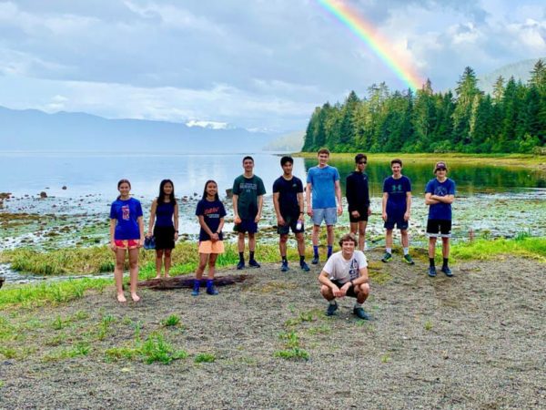 About ten runners in shorts and tshirts pose in front of a bay with a rainbow in the background
