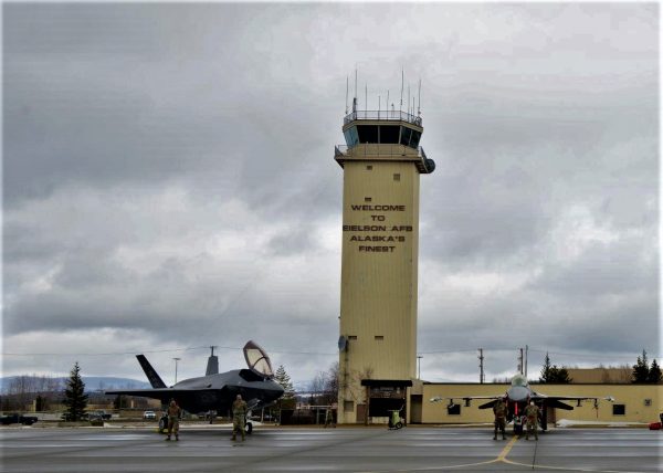 Two f-35 fighter jets are parked in front of a beige aircraft control tower