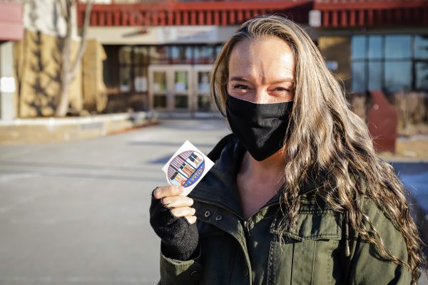 A white woman with a black facemask and blond hair holds an "I voted sticker: