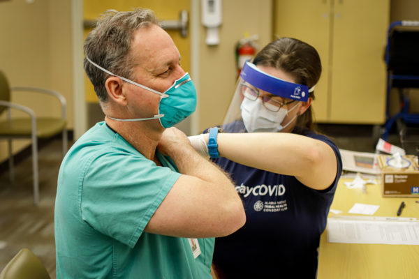 A man in green scrubs and a green n95 mask lifts up his shirt sleeve as a woman ina blue tshirt admdinisters a vaccine