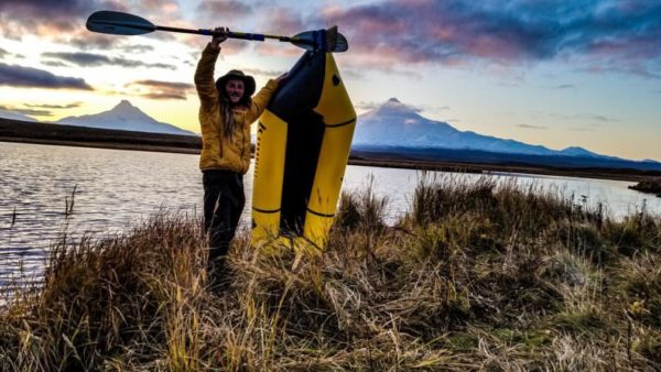 A hiker in a dry suit holds a padde and a packraft on an island. 