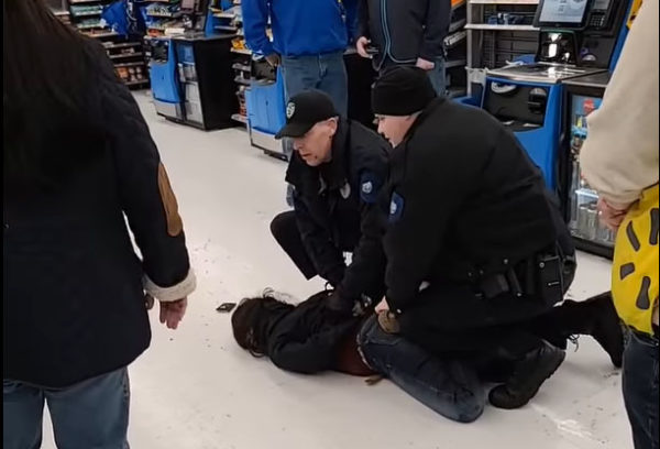 Two uniformed police officer crouch over a woman they have pinned to the floor in front of several grocery store checkout aisles.