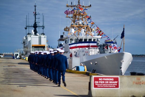 A white boat on a dock with a line of soldiers marching toward it