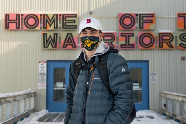 A student wearing a face mask, a baseball hat and a backpack stands in front of a school building.