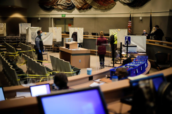an election official stands at a distance to a few people who are voting. other election officials and computers in the foreground