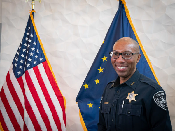 A black man in a police uniform smiles at an Alaska and U.S. flag