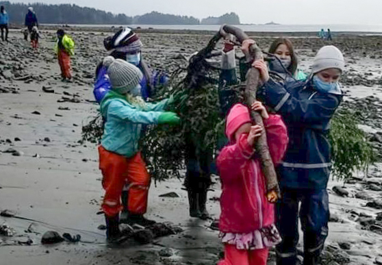 Kids in rain gear carry a spruec bough along the beach