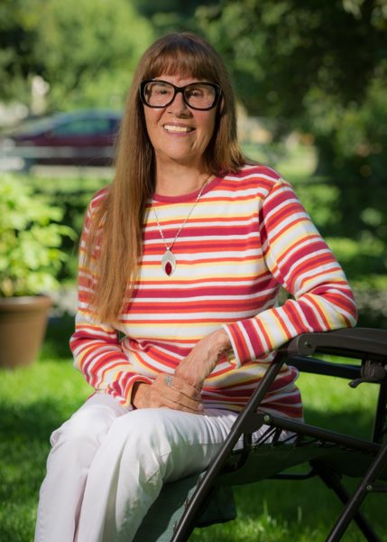 An older white woman wearing glasses and a striped shirt sits outdoors.