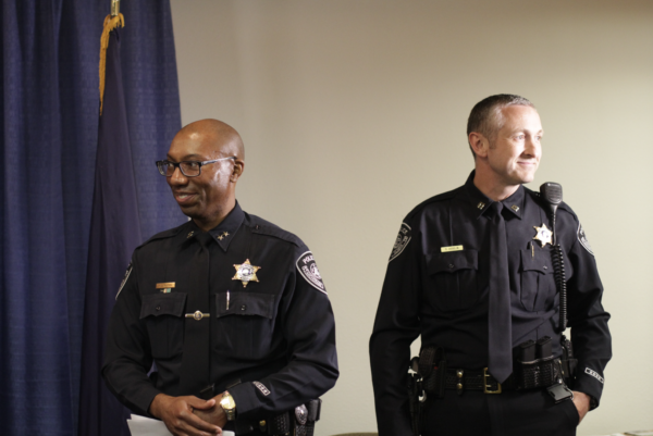 A black man in a police uniform and dark rimmed glasses smiles and a white man in a police uniform lookiing to the viewers right smiles