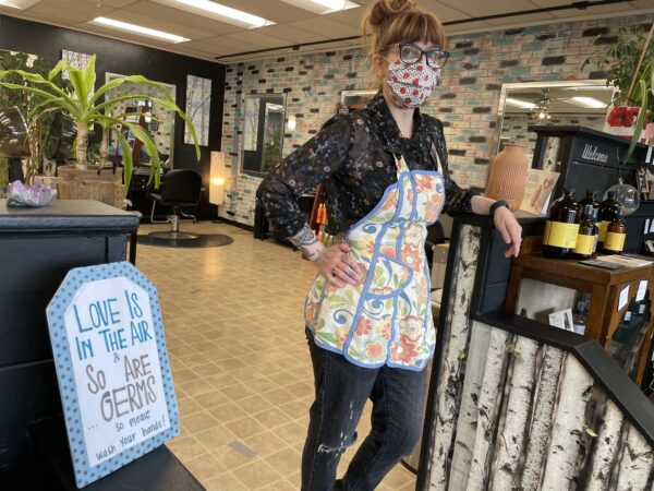 Masked lady in an apron stand in a salon. Sign in foreground reads "love is in the are and so are germs so please wash your hands."