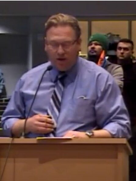 A white man in a purple shirt speaks at a podium