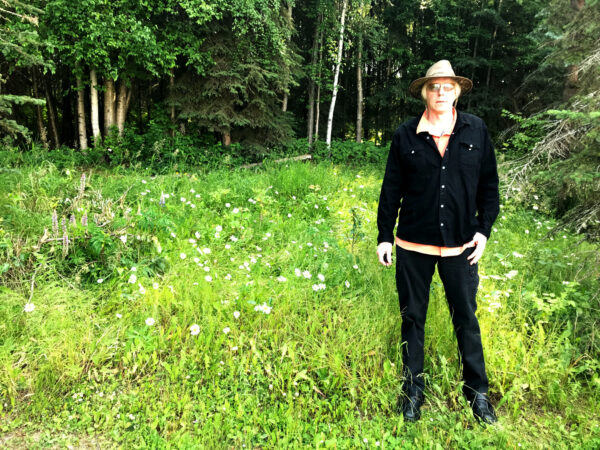 A man with light hair and a light hat, dark long sleeve shirt and dark pants stands on grass in front of trees.