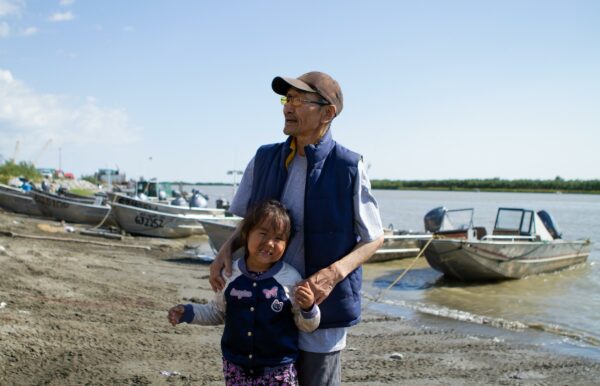 A man and a girl stand on a river shore with boats in the background.