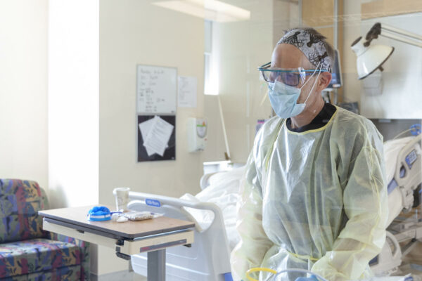 A woman sits in a hospital room wearing a face mask, face shield and gown.
