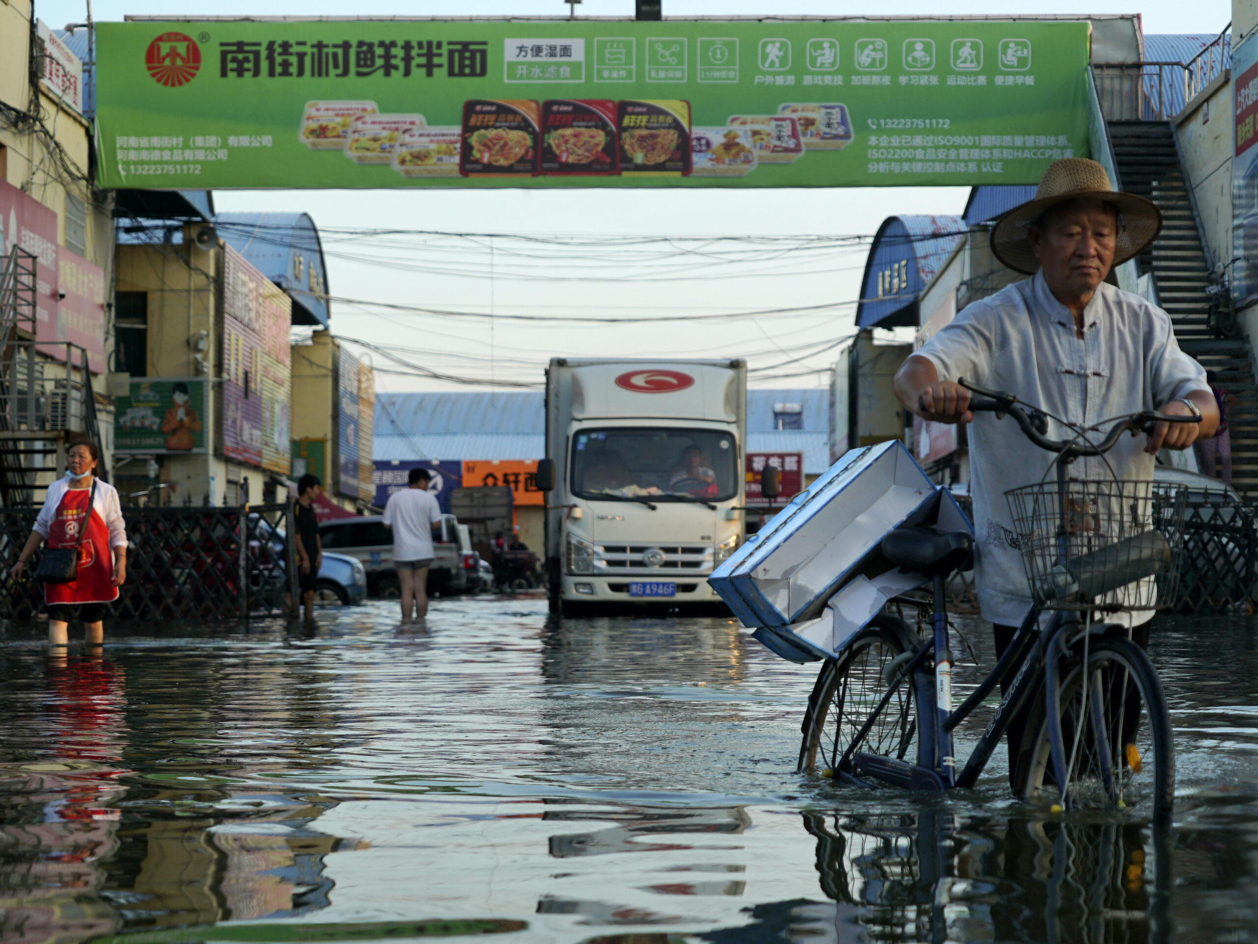 A truck tries to drive through a flooded street as other people walk and one man pushes a bicycle.