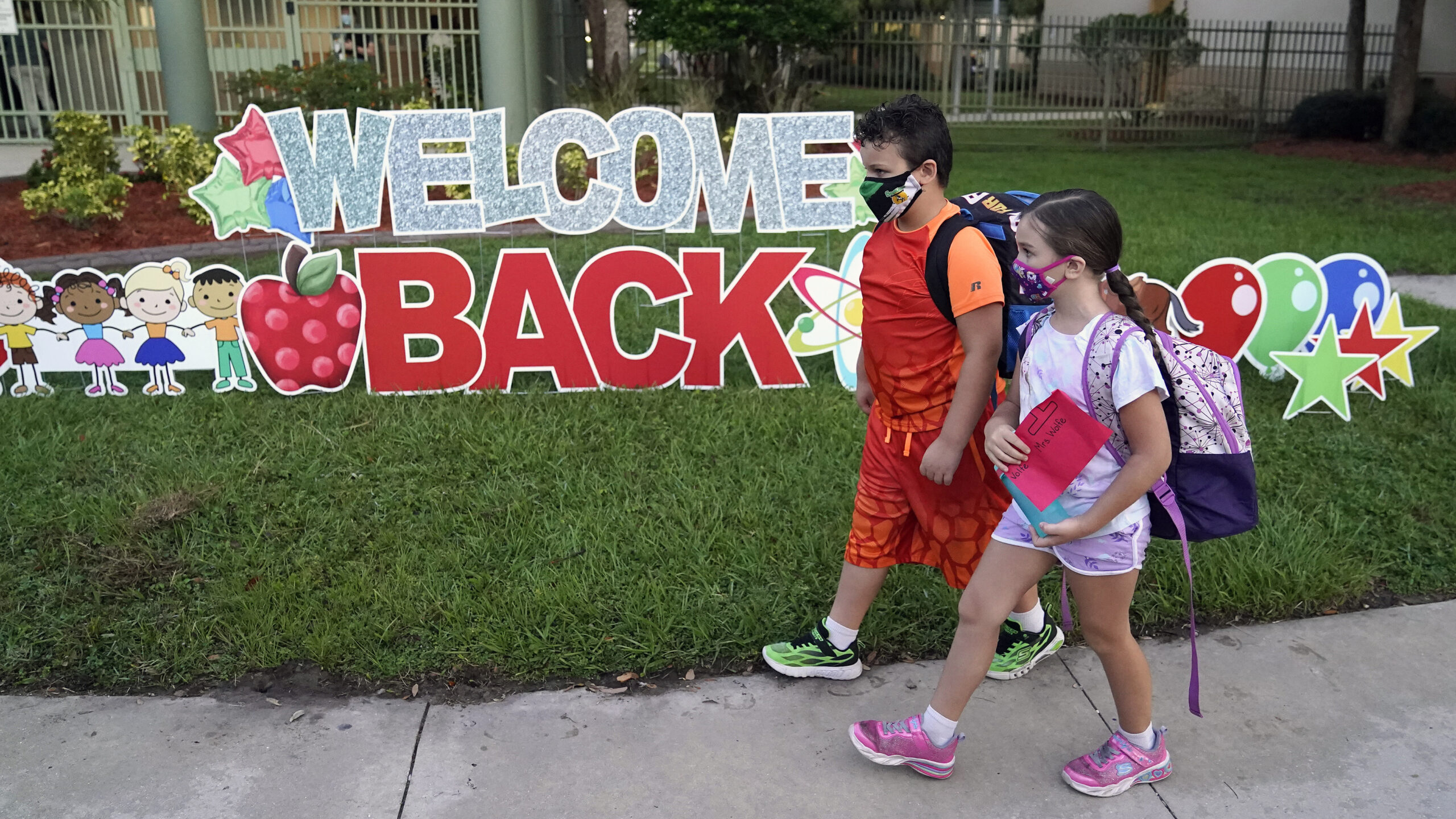 Two children walk on the sidewalk in face masks, on the grass is a big sign that says "Welcome Back"