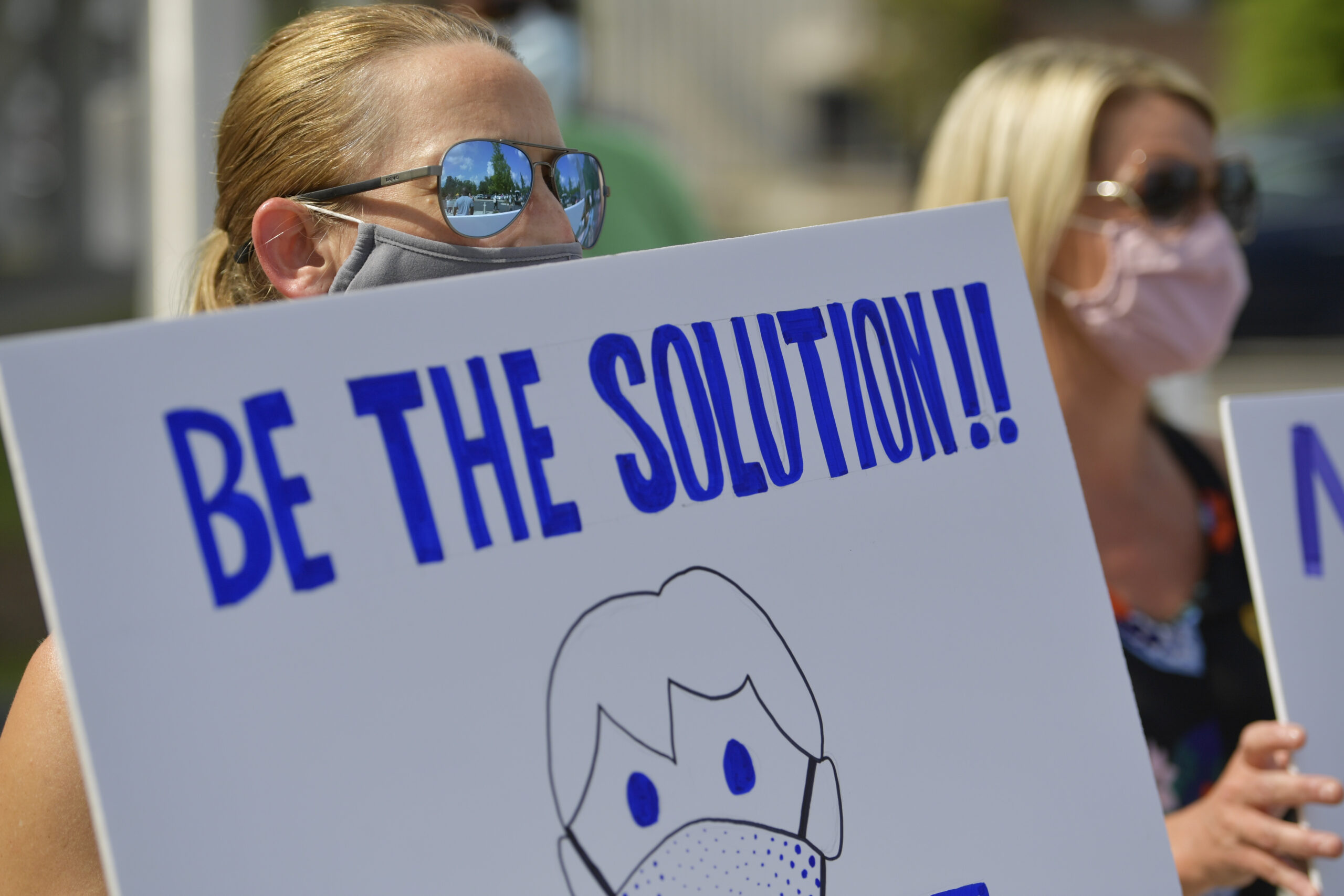 A woman in sunglasses and a mask holds a sign that says "Be the solution!" 