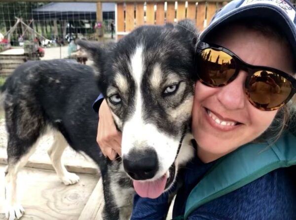 A woman in sunglasses and wearing a hat has her arm around a sled dog.