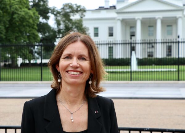 A woman facing the camera and smiling, while standing in front of the White House.