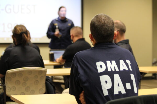 A person with a shirt that says DMAT PA-1 sits at a desk looking at a presenor at the front of the room
