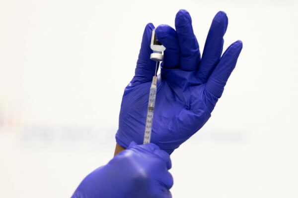 Hands in purple gloves pull a dose of vaccine from a vial.