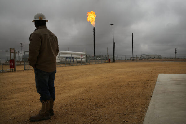 A person in a hard hat and jacket looks into the distance at fire coming from a tall cylinder.