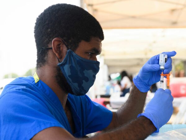 A health care worker in blue scrubs and wearing a mask prepares a dose of vaccine.