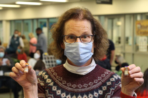 A woman in a face masks holds up two vials of vaccine.