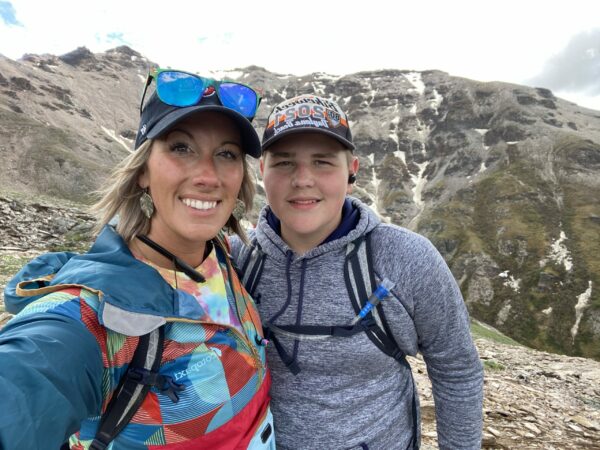 A white woman and a white teenage boy wearing hiking clothes and smiling on a mountain ridgeline.