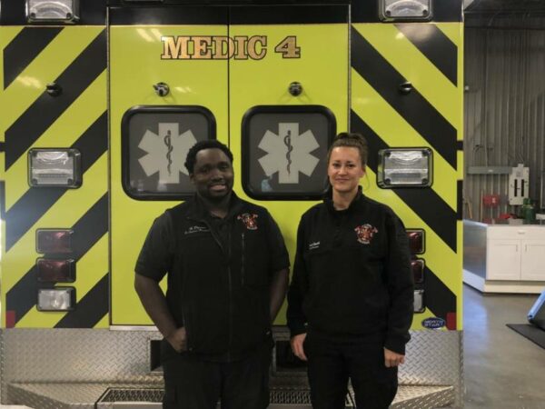 Two paramedics, in uniform, stand in front of the back of an ambulence.