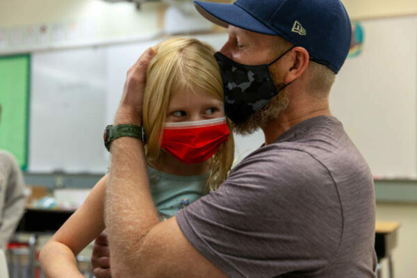 W white man with a t shirt and mask and baseball cap hugs a girl wearing a red face mask with blond hair