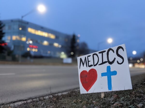 A white sign that says "medics" with a heart and blue cross painted below it, pictured in front of a road and a hospital behind it