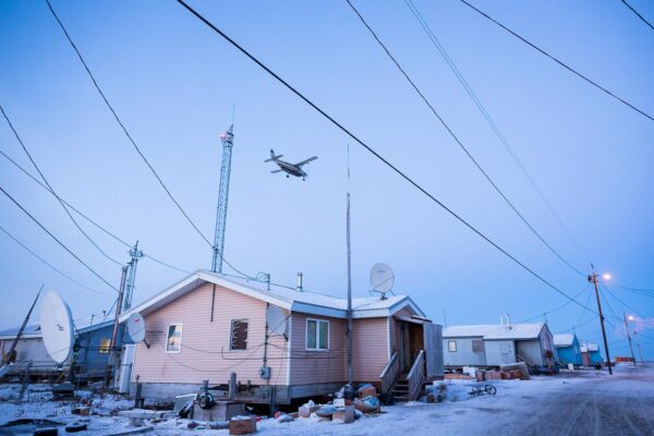 A pink house in a snowy road with a small plane flying above it and a lot of power lines hanging in between houses