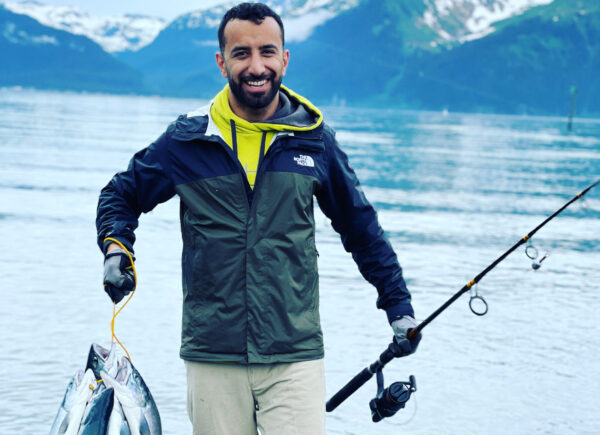 man holds a bunch of salmon by a cord in one hand and and fishing rod in the other. Water and mountains in background.