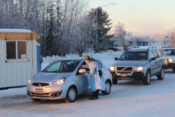 A person in a blue hospital gown reaches in the driver's window of a silver hatchback in a snowy parking lot next to a conex