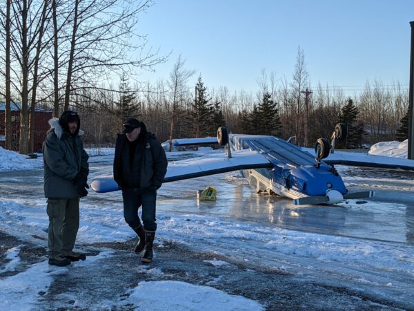 Two men walk on ice near a flipped-over small plane.