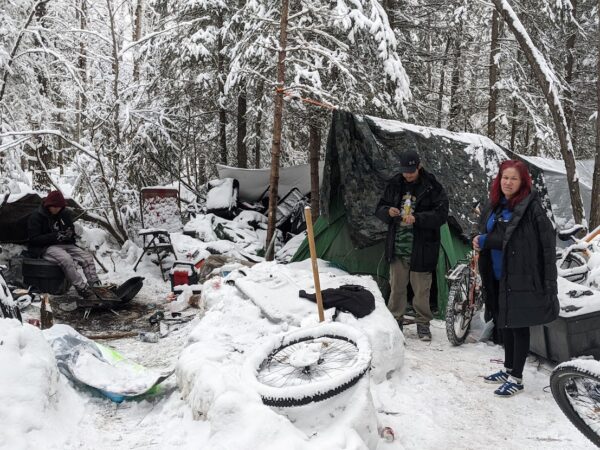 A woman with a coat draped around her and red hair next to a tent covered in a tar in a snowy forest. A man rolls a cigarrette next to a tent and another man sits by a fire pit. 