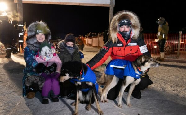 A family in coats with fur ruffs poses with sled dogs