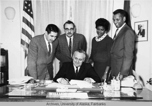 Black and white photo of Gov. Egan sitting at his desk signing a bill, with four people watching