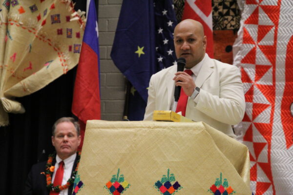 A polynesian man in a white suit jacket speaks into a microphone on a podium in front of some banners