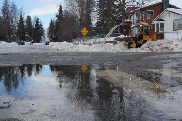 A snow blower crosses an intersection in front of a giant puddle in the roadway