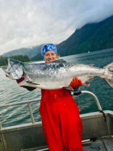 Seorang wanita muda berdiri di dek kapal memegang salmon raja yang sangat besar
