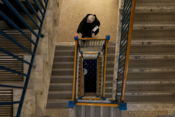 A woman in a mask standing on a landing, seen looking down through the stairwell from the landing below